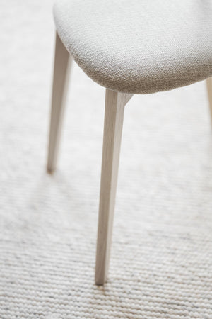 Närbild på stolsben i ljust trä och stoppad sits med beige/vit klädsel på vävd matta