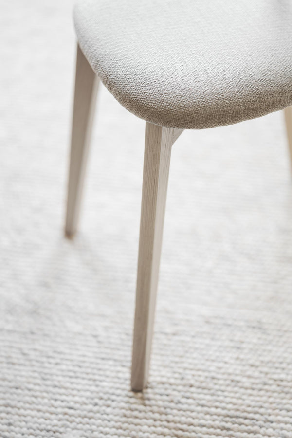 Närbild på stolsben i ljust trä och stoppad sits med beige/vit klädsel på vävd matta