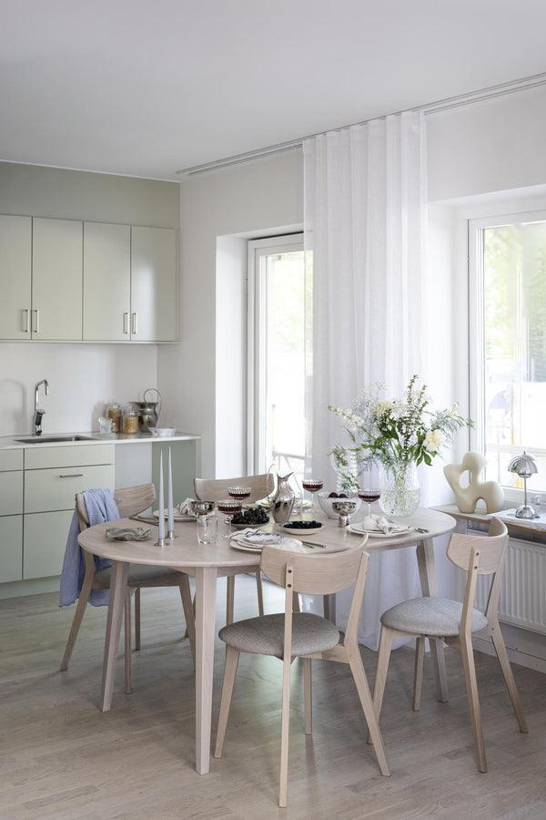 Ljust kök med runt matbord i ljust trä och matchande stolar med grå dynor, vita skåp och gardin