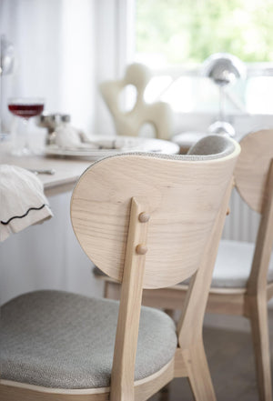Ljus trästol med rundad rygg och grå dyna vid matbord med dukning i ljust kök