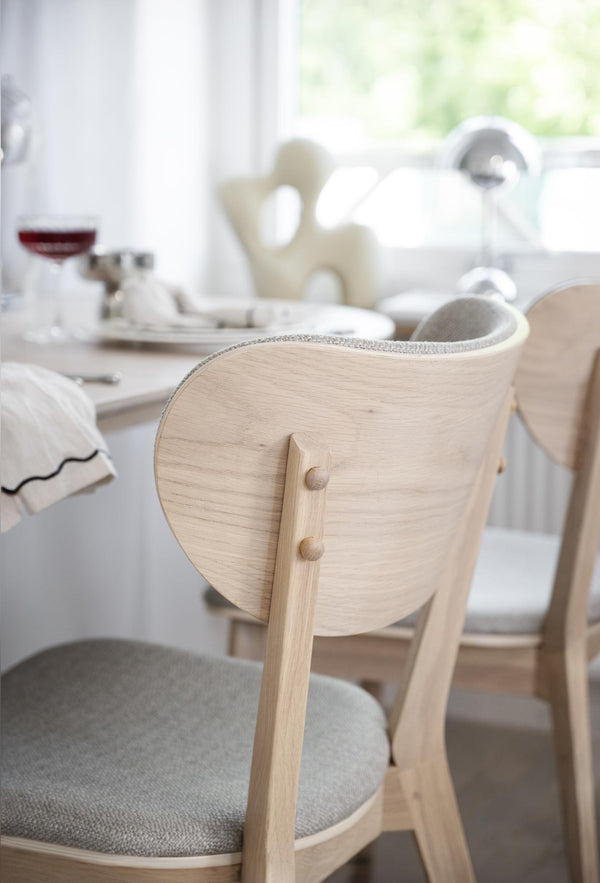 Ljus trästol med rundad rygg och grå dyna vid matbord med dukning i ljust kök