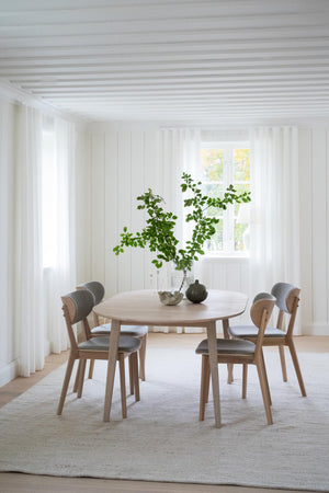 Runt matbord i ljust trä med fyra stolar och grå dynor i minimalistisk matsal med vita väggar