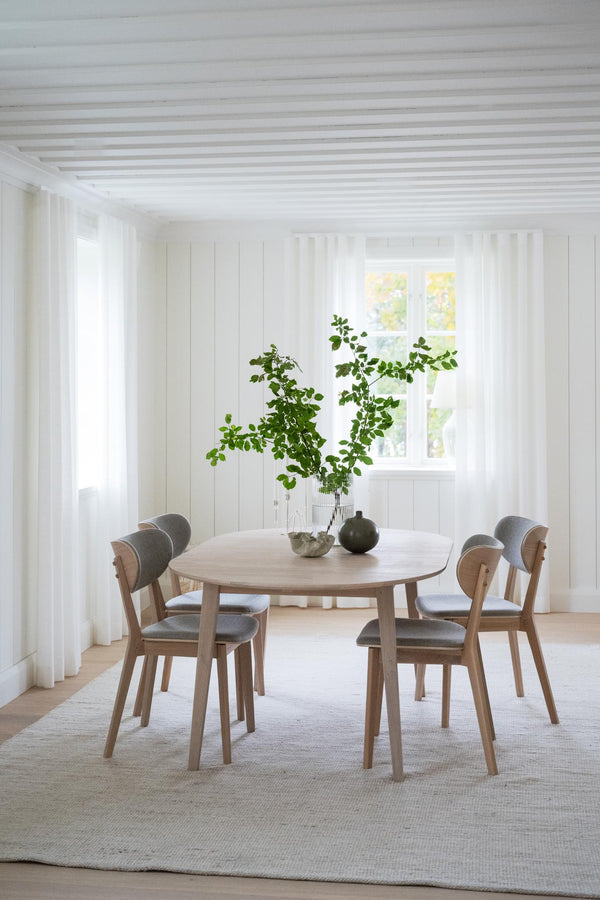 Runt matbord i ljust trä med fyra stolar och grå dynor i minimalistisk matsal med vita väggar