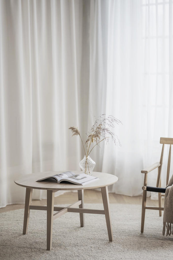 Ljust vardagsrum med runt soffbord i trä, tidning, vas med torkade gräs och minimalistisk stol mot vita gardiner