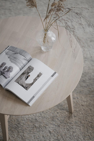 Ljust träbord i rund form med öppen bok och glasvas med torkade gräs på vävd matta