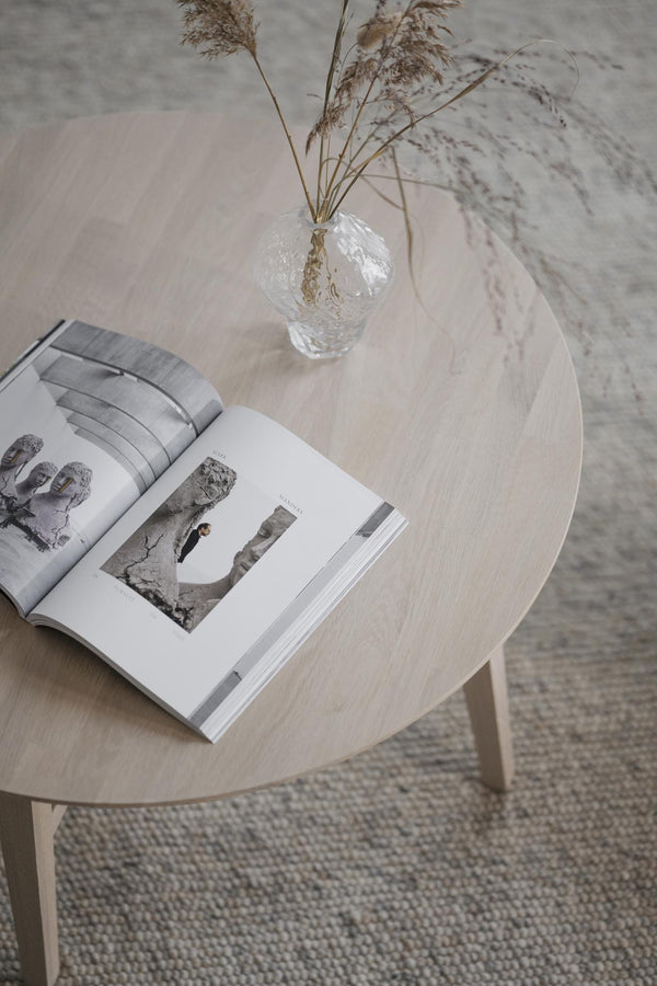 Ljust träbord i rund form med öppen bok och glasvas med torkade gräs på vävd matta