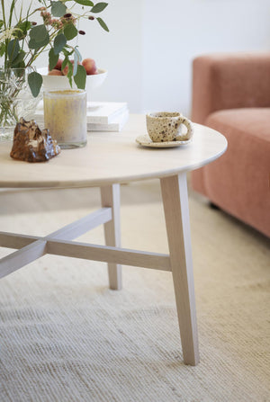 Runt soffbord i ljust trä med vita ben, dekorerat med spräcklig keramikmugg, vas och ljus på matta
