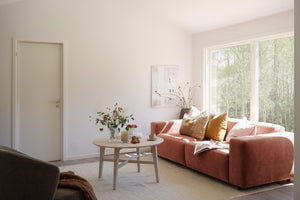 Ljust vardagsrum med korallrosa soffa, runda soffbord i trä och stora fönster mot grön skog