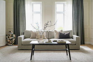 Elegant vardagsrum med beige soffa, svartvitt soffbord, gröna gardiner och dekorativa kuddar vid fönster