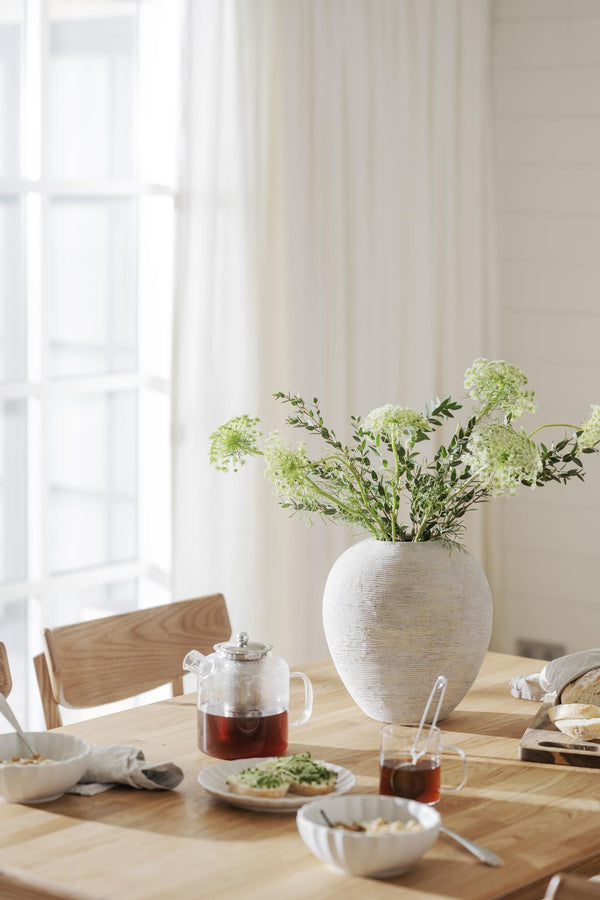 Ljus matplats med vit keramisk vas fylld med gröna blommor, tekanна och frukost på träbord