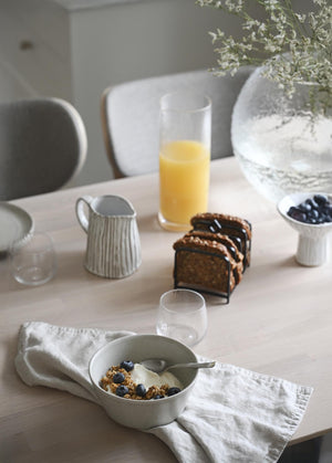 Frukostbord med granola i skål, blåbär, juice, bröd och vita porslinskärl i minimalistisk stil