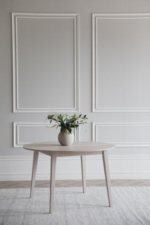 Runt matbord i ljust trä med vas och blommor, placerat på vit matta mot vägg med vit inramning