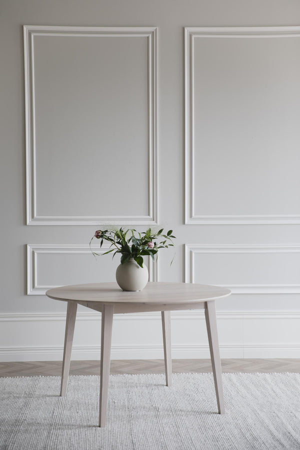 Runt matbord i ljust trä med vas och blommor, placerat på vit matta mot vägg med vit inramning