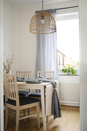 Taklampa i flätad rotting hänger över matbord i ljus matsal med vita stolar och grå gardiner
