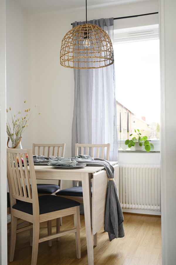 Taklampa i flätad rotting hänger över matbord i ljus matsal med vita stolar och grå gardiner