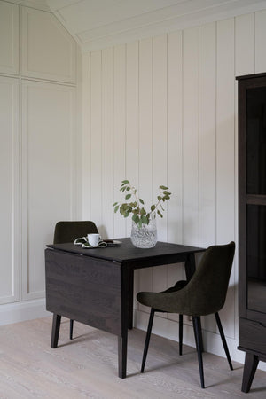 Svart skrivbord med två gröna stolar vid vit träpanel, dekorerat med kaffekopp och växt i glasvas