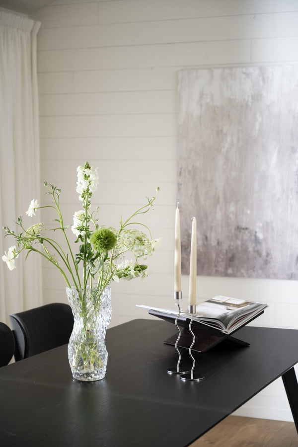 Svarta ljusstakar i vågformat design med vita ljus på matsalsbord bredvid kristallvas med vita blommor