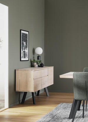 Skänk i ljust trä med svarta ben i modern skandinavisk stil, dekorerad med lampa och växt mot grön vägg