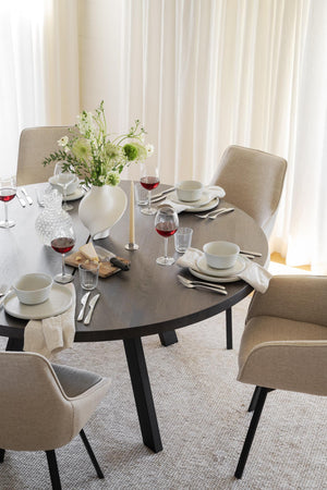 Rund matbord i mörkgrått med beige stoppade stolar, uppdukat med vinglas, tallrikar och blomvas med vita blommor