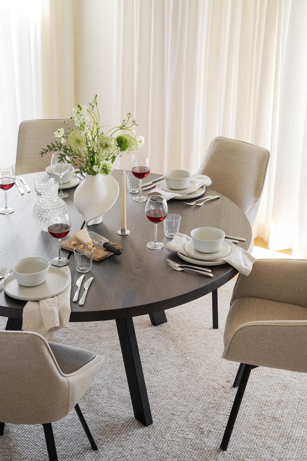 Rund matbord i mörkgrått med beige stoppade stolar, uppdukat med vinglas, tallrikar och blomvas med vita blommor