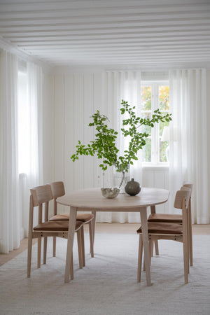 Runt matbord i ljust trä med fyra stolar i minimalistisk skandinavisk matsal med vita väggar och gardiner