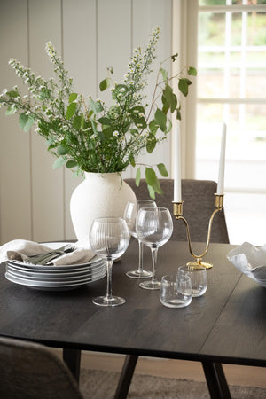 Elegant dukat matbord med vit vas, gröna blommor, vinglas, tallrikar och guldljusstake i modern skandinavisk stil