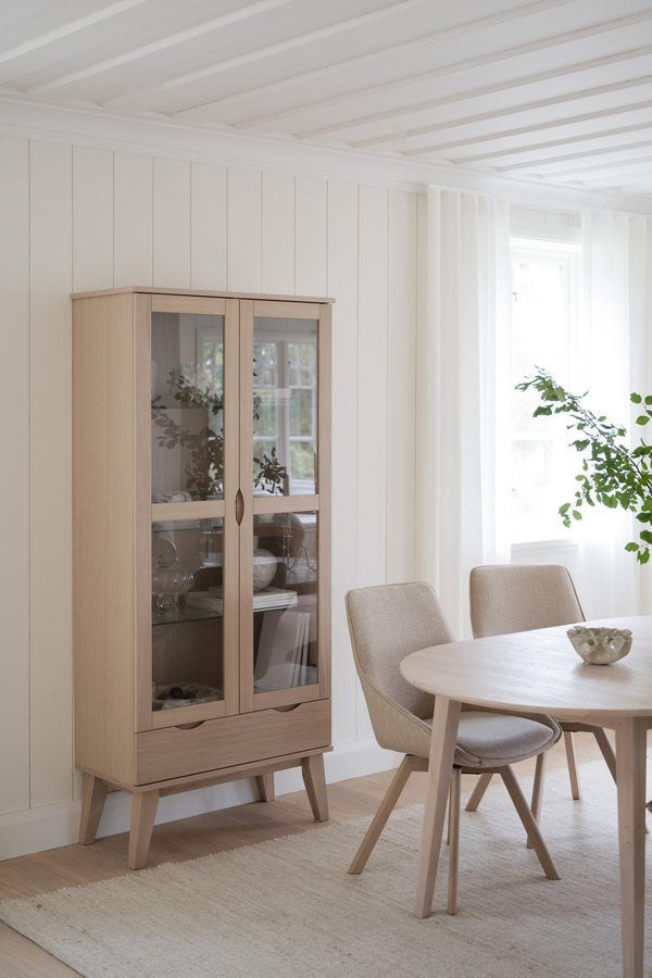 Vitrinskåp i ljust trä med glasdörrar bredvid matbord och stolar i skandinavisk stil
