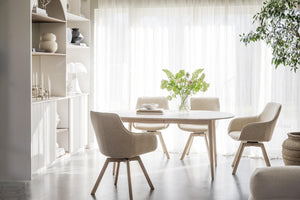 Ljus matplats med runt matbord i trä och beige stolar mot fönster med vita gardiner