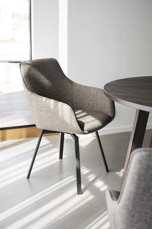 Grå matstol med armstöd och svarta ben bredvid runt matbord i modernt minimalistiskt rum med stora fönster