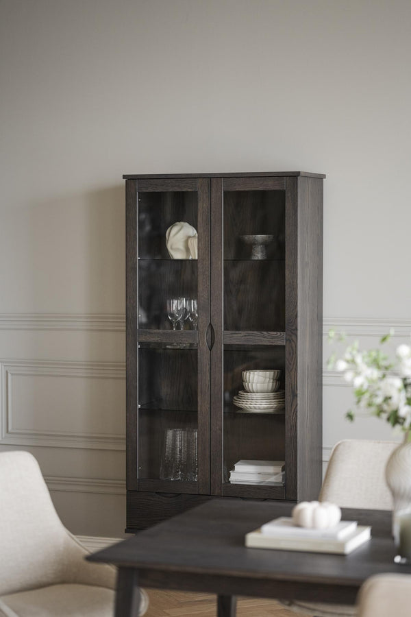 Vitrinskåp i mörkgrått trä med glasdörrar och hyllplan, inrett med porslin och dekorationer i vardagsrum