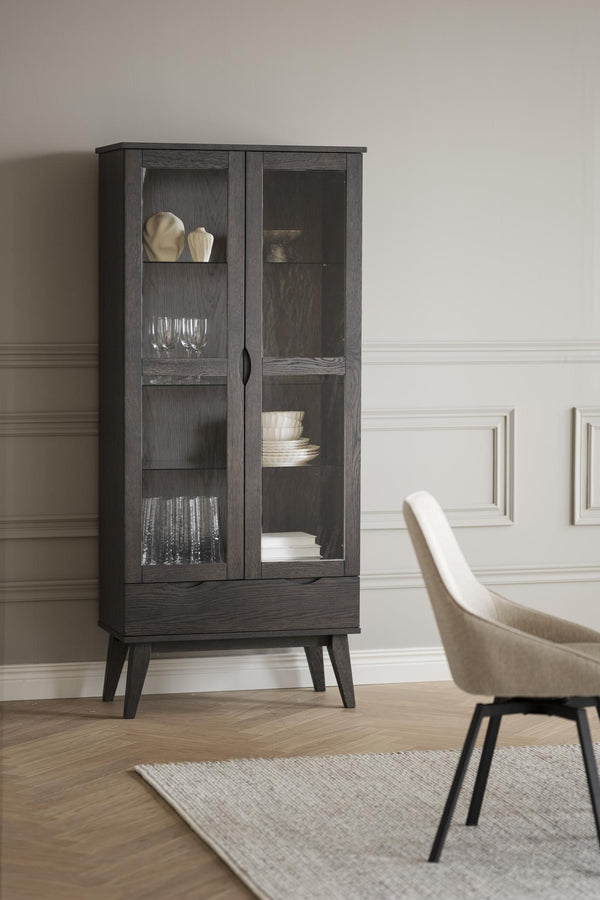 Mörkgrått vitrinskåp med glasdörrar och låda på träben, inrett med glas och porslin