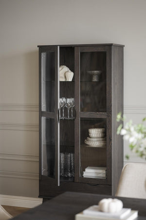 Mörkbrunt vitrinskåp med glasdörr och öppna hyllor, inrett med porslin, glas och dekorationer