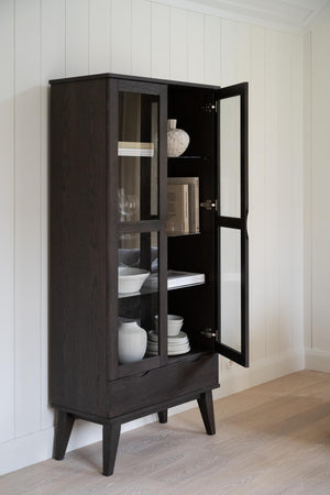 Mörkbrun vitrinskåp i trä med glasdörrar och synliga hyllor med porslin och dekorationer