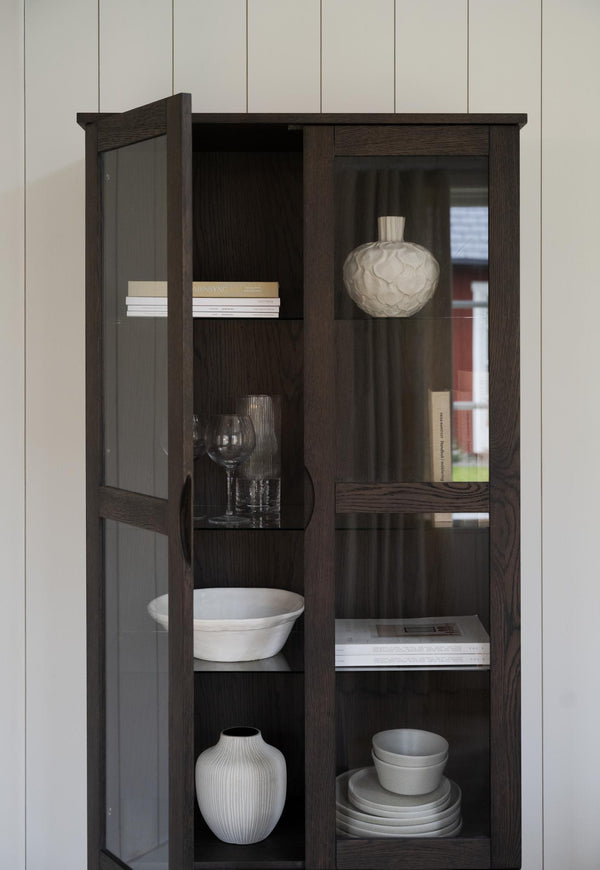 Mörkbrun vitrinskåp med glashylla och glasdörrar, inredd med vita vaser, tallrikar och böcker