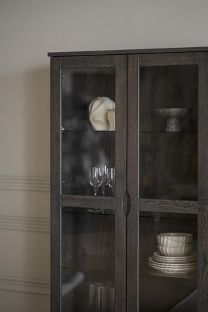Vitrinskåp i mörkt trä med glashyllor och diskret handtag, inrett med porslin och glasföremål