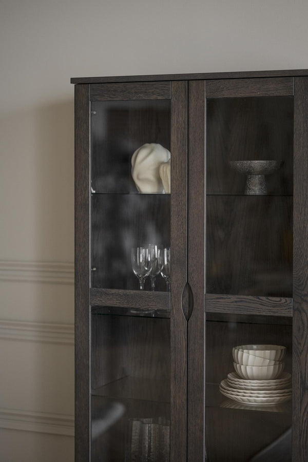 Vitrinskåp i mörkt trä med glashyllor och diskret handtag, inrett med porslin och glasföremål