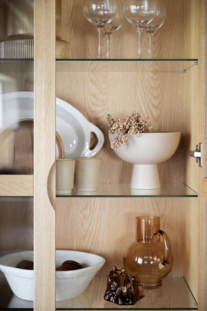 Ljust ekskåp med glashyllor och keramiska skålar, glas och dekorativa föremål i neutrala toner