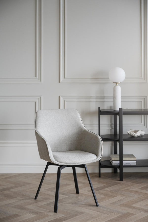 Ljusgrå stoppad stol med svarta ben i modern skandinavisk design framför vit panelvägg med hylla