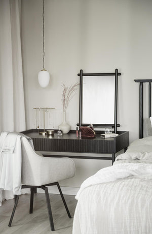 Minimalistiskt sovrum med mörkgrå sidobord, vit pendellampa, spegel och ljusgrå stol mot vit vägg