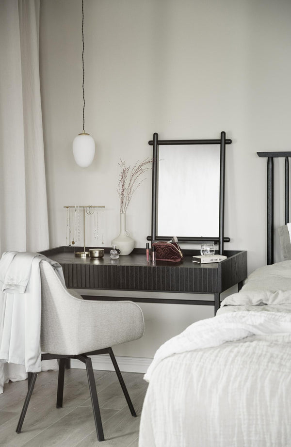 Minimalistiskt sovrum med mörkgrå sidobord, vit pendellampa, spegel och ljusgrå stol mot vit vägg