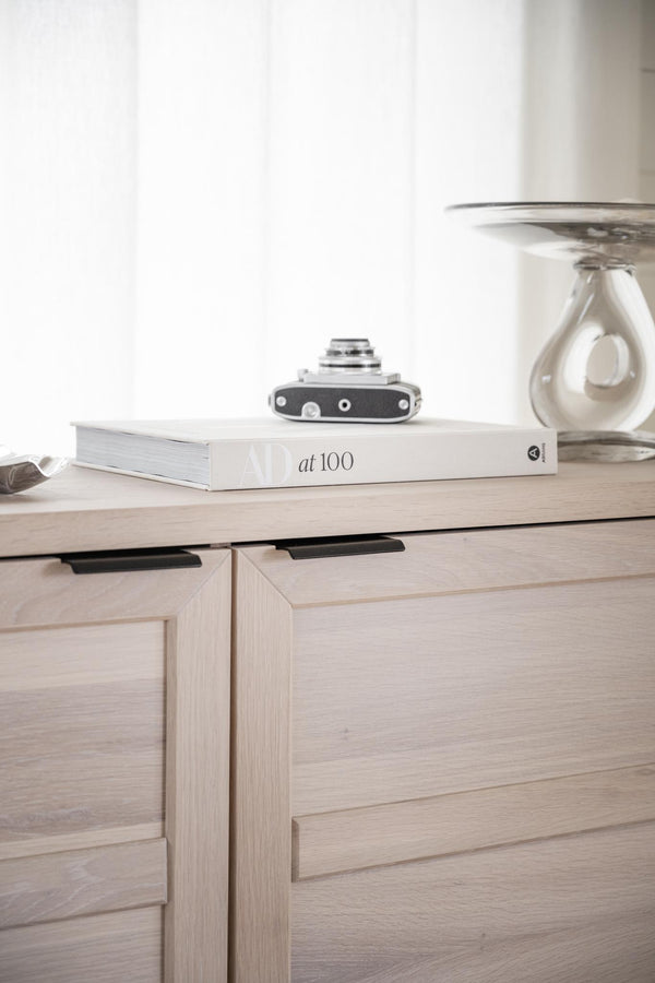 Ljust träskåp med lådor, bok märkt "at 100", vintage kamera och glasvas i minimalistisk inredning