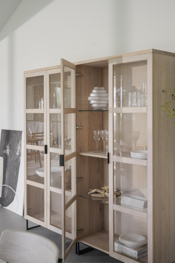 Vitrinskåp i ljust trä med glasdörrar, fyllda med glas, tallrikar och dekorativa föremål i minimalistisk stil