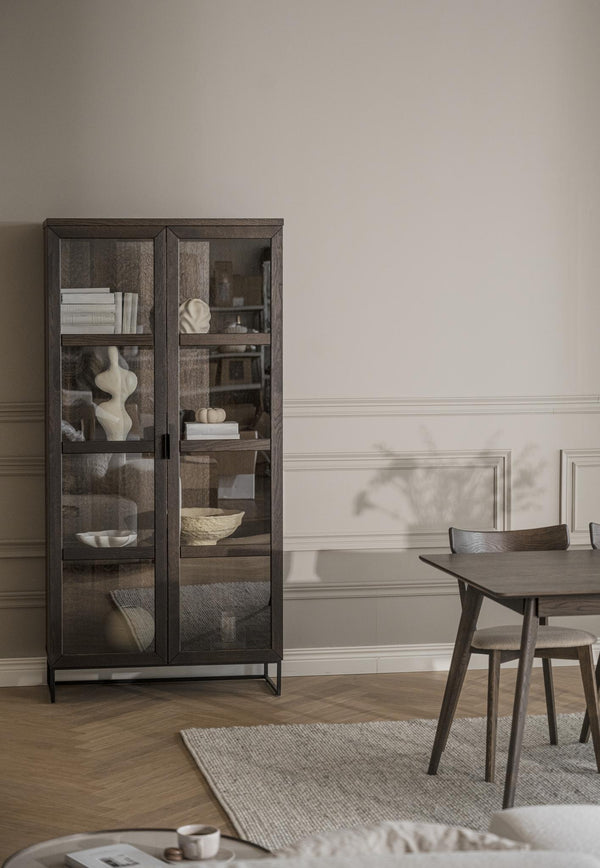 Vitrinskåp i mörkt trä med glashyllor och synliga föremål i elegant matsal med bord och stolar