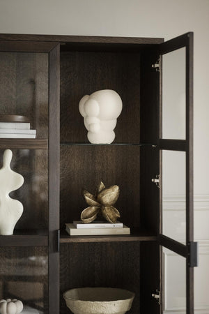 Mörkt trähylla med glaskiva, skulpturer i vitt och guld samt böcker bakom glasdörr