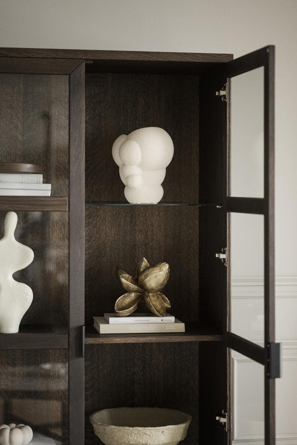 Mörkt trähylla med glaskiva, skulpturer i vitt och guld samt böcker bakom glasdörr