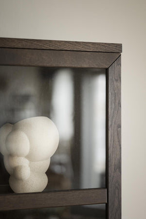 Dekorativ skulptur i ljus sten placerad i mörkbrun hylla med glasdörr