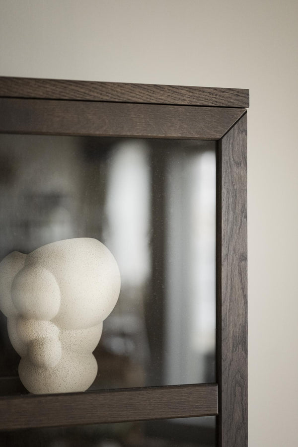 Dekorativ skulptur i ljus sten placerad i mörkbrun hylla med glasdörr