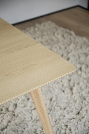 Närbild på ljust träbord med synlig trästruktur och ben, placerat på grå luggmatta i modern inredning