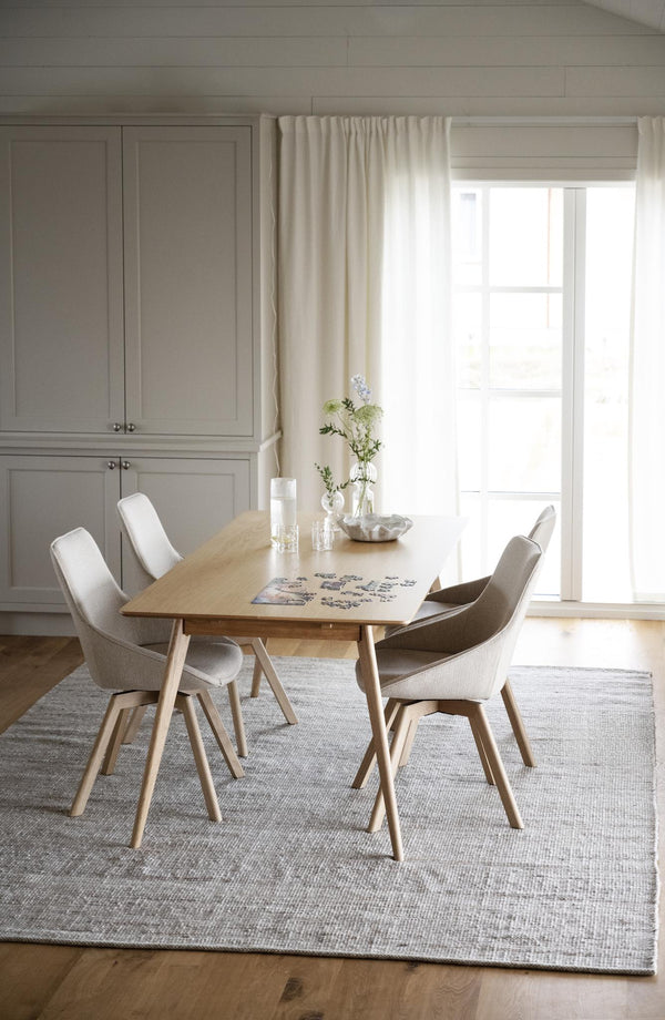 Ljus matplats med runt matbord i trä, fyra vita stolar och grå matta under fönster med gardiner