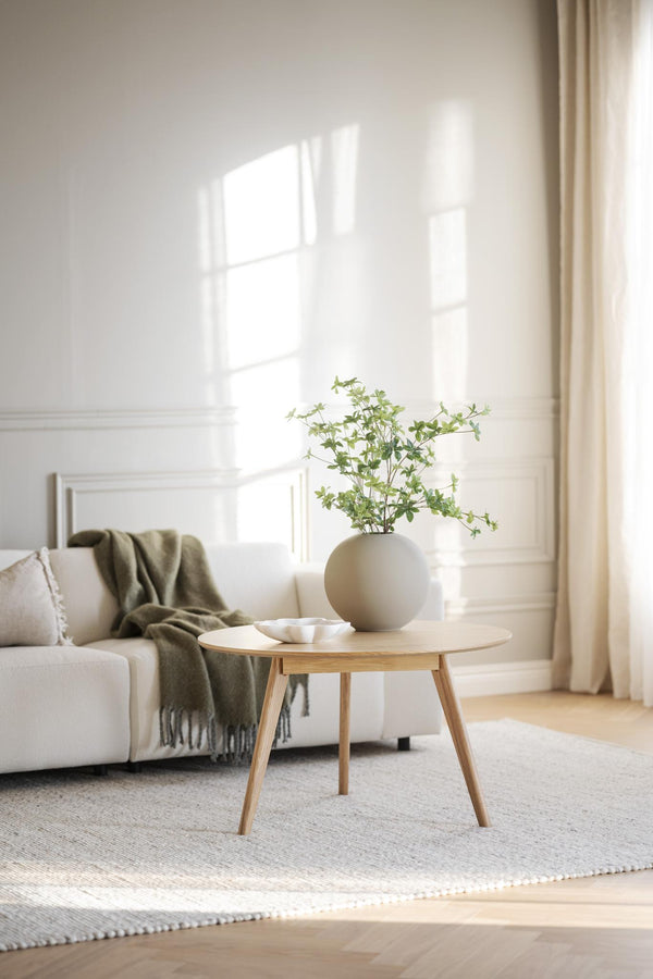 Ljust vardagsrum med beige soffa, runt träbord, grön filt och vas med gröna växter i minimalistisk stil